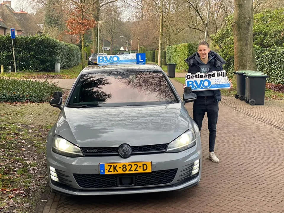 Geslaagde leerling met het bord Geslaagd bij BVO naast een lesauto van BVO in een straat met bomen