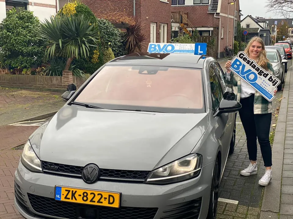 Geslaagde leerling houdt het bord Geslaagd bij BVO omhoog naast een lesauto van BVO